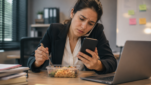 Mulher sentada à mesa durante o horário de almoço segura um celular enquanto olha mensagens de trabalho. À sua frente há uma marmita aberta e um notebook ligado, transmitindo a sensação de que o descanso foi interrompido por demandas profissionais.