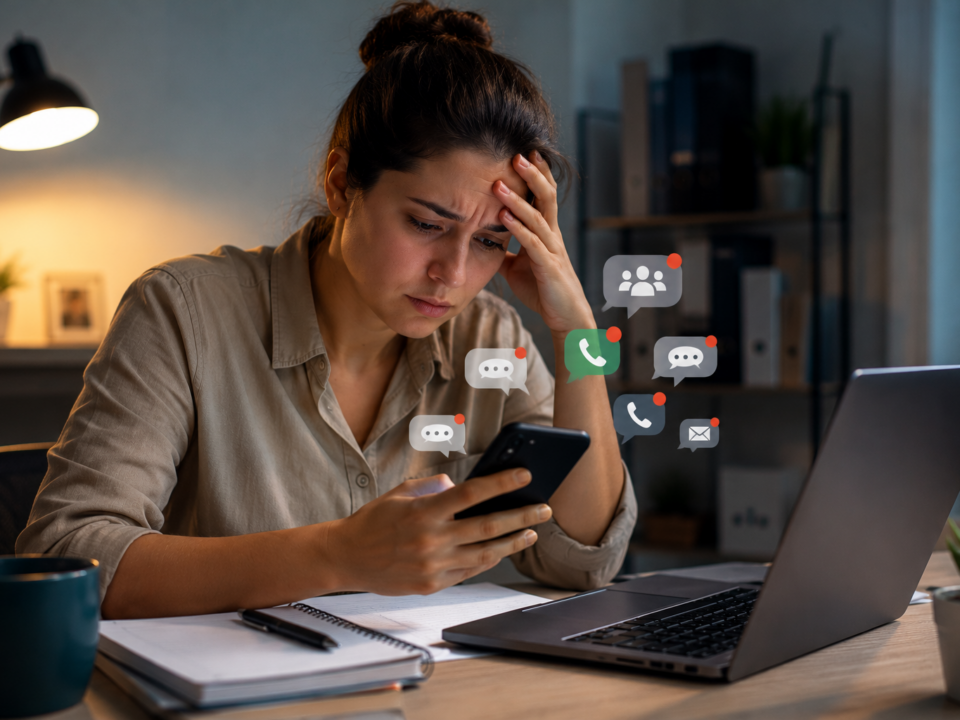 Mulher sentada em uma mesa de trabalho observa o celular com expressão de preocupação enquanto recebe diversas notificações relacionadas ao trabalho, mesmo fora do ambiente corporativo.