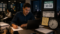 Homem continua trabalhando em um escritório à noite, mesmo após registrar a saída às 18h. Ao lado dele, um painel mostra o horário registrado e um relógio próximo das 19h, representando a realização de horas extras não registradas.